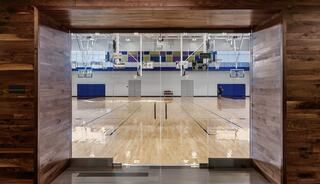 A modern gymnasium features a polished wooden floor, basketball hoops, and a spacious court, framed by a wooden entrance.