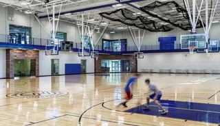 A spacious indoor basketball court features two players in motion, bright lighting, and a modern design with multiple hoops and seating areas.