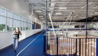 A person jogs along a blue indoor track overlooking a spacious gym with basketball courts and large windows allowing natural light.