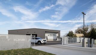 A modern gray building with a fenced entrance, surrounded by greenery and a clear sky. A car is parked in front, creating a welcoming atmosphere.