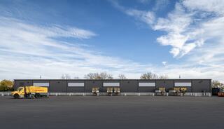 A row of large gray warehouses with loading docks and parked yellow trucks under a bright sky, showcasing an industrial setting.