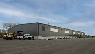 A large, modern warehouse with a gray exterior, loading dock, and parked trucks, set against a cloudy sky.