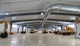 A spacious industrial warehouse features various construction equipment, with high ceilings and large windows allowing natural light.