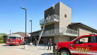 Firefighters practice on a training tower with a ladder, while fire trucks are stationed nearby at a fire station. Bright sunny day.