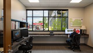 A modern office with two workstations, one person at a desk, and a large window showing a fire truck outside. Bright, functional space.