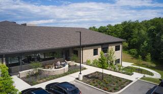 A modern building with a landscaped entrance and outdoor seating, surrounded by greenery and a parking lot under a clear sky.