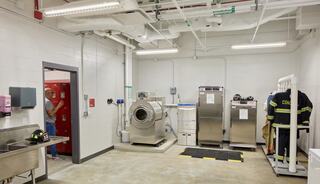 A clean, well-lit laundry room features a large washer, dryers, lockers, and firefighter gear hanging on a rack.