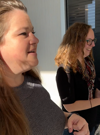 Two women are chatting in bright light, smiling and engaged in conversation. One has long, straight hair, while the other has curly hair and glasses.