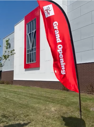 A vibrant red flag announcing "Grand Opening" stands in front of a modern building with large windows and a decorative "V" design.