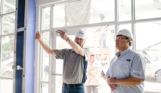 Two men in hard hats discuss a project inside a building under construction, with additional workers visible in the background.