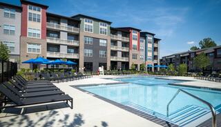 A modern apartment complex features a sparkling pool surrounded by lounge chairs and vibrant blue umbrellas on a sunny day.