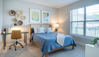 A cozy bedroom features a blue bedspread, modern art, a lamp, a desk, and large windows allowing natural light to brighten the space.