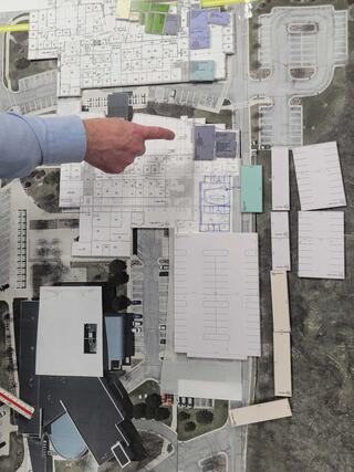 A hand points to a detailed layout map, featuring buildings, parking areas, and surrounding landscapes, illustrating a planned development site.