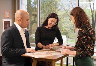 Three people collaborate at a table, discussing documents and sharing ideas in a bright, modern space with a view of nature outside.