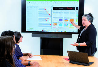 A woman presents data on a screen to a group, discussing charts and information displayed, while a laptop is open on the table.