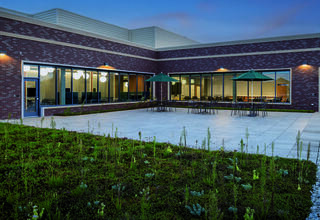 A modern courtyard features stone flooring, outdoor seating under green umbrellas, and lush, low-maintenance landscaping, set against a brick building.