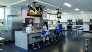 A modern kitchen with a large island, equipped with cooking stations and screens displaying culinary demonstrations, hosts two seated learners.