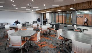 A modern, spacious room features round tables with chairs, cozy seating areas, and large windows. People are engaged in conversation and movement.