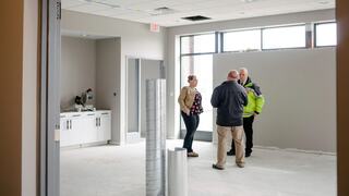 Three people are discussing in an empty, partially finished room with natural light, featuring gray walls and construction materials scattered around.