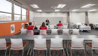 A modern classroom setup with students in red shirts engaging with an instructor at a front podium, featuring medical equipment in the background.