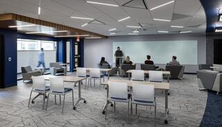 A modern classroom features flexible seating and a presentation area, with students engaged in discussion and a lecturer at the front.