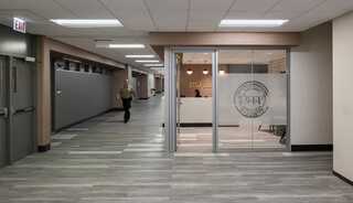 A modern office space with glass walls, warm lighting, and a spacious hallway featuring a person walking in the background.