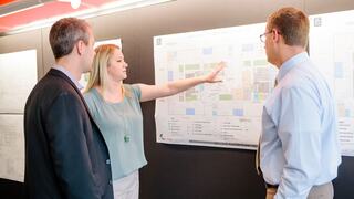 A group discusses plans, with a woman pointing to a map while two men listen attentively, showcasing collaboration and strategy.