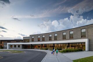 A modern brick building with large windows under a cloudy sky, featuring two children walking towards the entrance, surrounded by greenery.