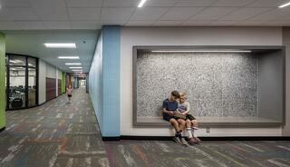 A colorful school hallway features two children sitting together in a cozy nook, while another child walks in the distance.