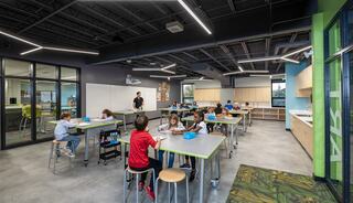A bright, modern classroom with students engaged at tables, a teacher guiding them, and a spacious layout featuring interactive learning areas.