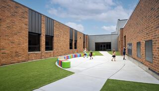 A play area features colorful seating and children engaging in activities on a well-maintained lawn, surrounded by brick buildings under a blue sky.