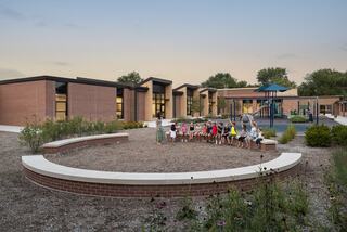 A playground area with children gathered in a semi-circle, surrounded by modern brick buildings and greenery at dusk.