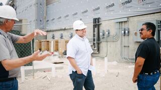 Three men in hard hats converse on a construction site, with building materials and electrical connections visible in the background.