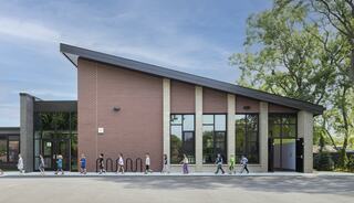 A modern school building features a unique sloped roof and large windows, with a line of students walking along the pathway. Lush greenery surrounds.