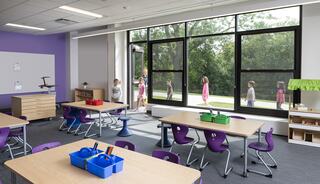 A bright, modern classroom features colorful furniture and a view of children walking outside, enhancing the learning environment.