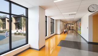 A spacious school hallway features large windows, vibrant flooring, and colorful artwork, with students walking toward classrooms.