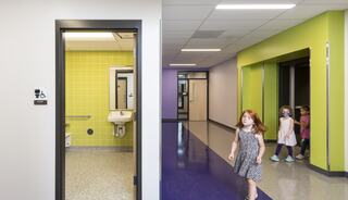 A colorful school hallway features a girls' restroom with bright yellow tiles, while children wander about, enjoying their environment.