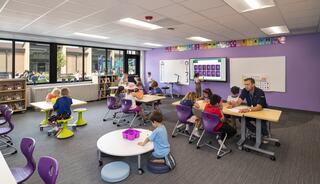 A bright classroom filled with children engaged in activities, featuring modern furniture and a large screen displaying educational content.