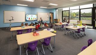 A colorful classroom features children sitting in a circle, attentive to a teacher. Bright furniture and large windows create a welcoming atmosphere.