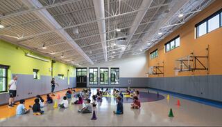 A group of children sits on the gym floor, arranged around colorful cones, while an instructor leads a session. Brightly lit and welcoming space.