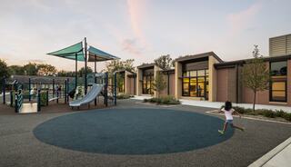A playground with slides and climbing structures is bathed in warm evening light, while a child runs joyfully on soft rubber ground.