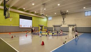 A vibrant gymnasium filled with children playing and engaging in activities, highlighted by a large screen and colorful cones scattered on the floor.