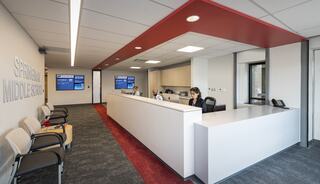A modern office interior featuring a reception area, staff at desks, and informational screens, designed for a middle school environment.