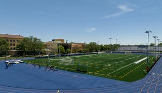 A spacious athletic field features bright turf, running tracks, and soccer goals, surrounded by trees and modern buildings under a clear blue sky.
