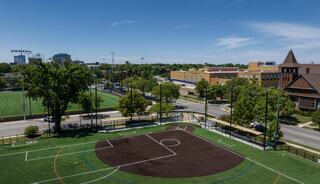A vibrant sports field with green turf and marked lines, surrounded by trees and urban buildings under a clear blue sky.
