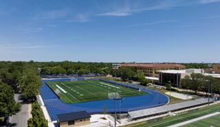 A bright, open athletic field with blue track and grass, surrounded by trees and school buildings under a clear sky.