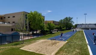 A sunny outdoor track and field area features lanes, a long jump pit, and athletes training, surrounded by green trees and a sports facility.