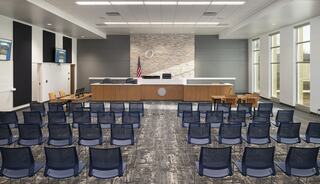 A modern courtroom featuring a wooden bench, chairs arranged for seating, large windows, and a backdrop with an emblem.