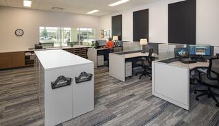 A modern office space with gray carpet, sleek white desks, and two workstations. Natural light enters through large windows.
