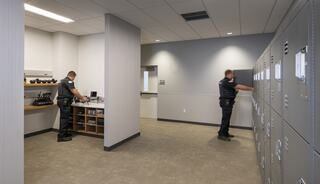 Two officers are in a modern locker room, one organizing equipment on a counter and the other using a locker.